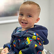 Brody joined the competition — help win amazing prizes! blurred_background, bright, casual, child, clothing, colorful, cute, face, happy, indoors, jacket, pattern, portrait, seat, short_hair, smiling, toddler, warm, window, young_child