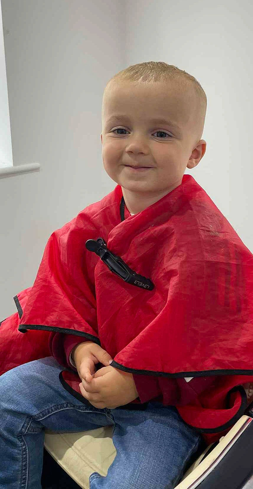 blond_hair, boy, casual_clothing, chair, child, clasped_hands, cute, haircut, happy, indoor, jeans, light_skin, minimalist, portrait, red_cape, sitting, smiling, toddler, window, young