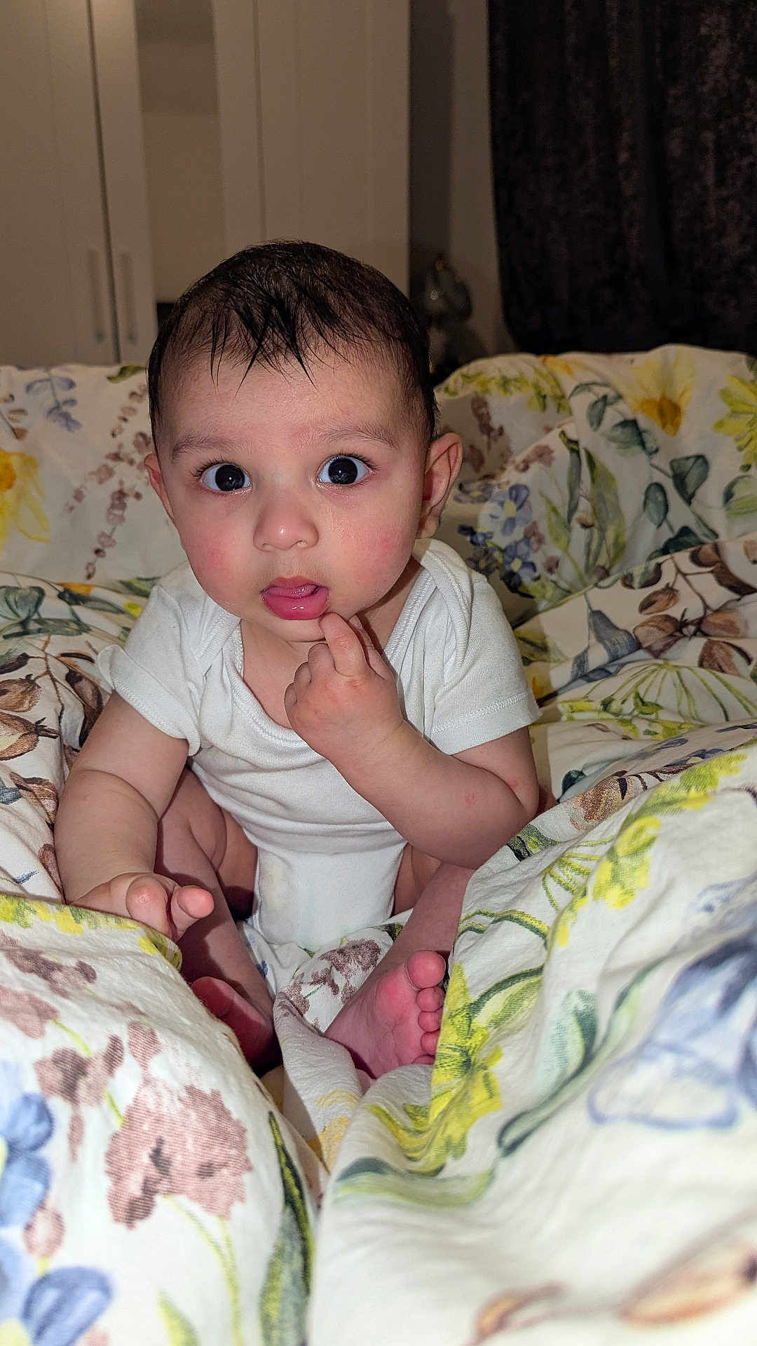 Harvin is registered to the contest to win money with this photo: baby, infant, child, face, eyes, hand, finger, mouth, white_onesie, blanket, floral_pattern, bedroom, indoors, curious, sitting, skin, foot, expression, person, portrait