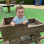 toddler, child, toy_car, wooden_toy, playground, green_surface, bench, bicycle, book_kiosk, outdoor, smiling, tongue_out, bib, casual_clothing, curly_hair, daylight, play, fun, wood, nature
