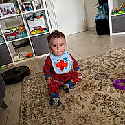 Kailan a rejoint le concours — aidez-le/la à gagner de superbes lots ! baby, bib, carpet, child, clothing, curious, door, face, floor, home, indoor, living_room, person, photograph, red_pajamas, rug, shelf, sitting, toddler, toy