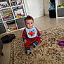 baby, bib, carpet, child, clothing, curious, door, face, floor, home, indoor, living_room, person, photograph, red_pajamas, rug, shelf, sitting, toddler, toy