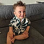 toddler, child, smiling, sitting, couch, gray, shirt, patterned_clothing, hair, happy, indoors, face, person, barefoot, legs, hands, glasses, wall, texture, casual