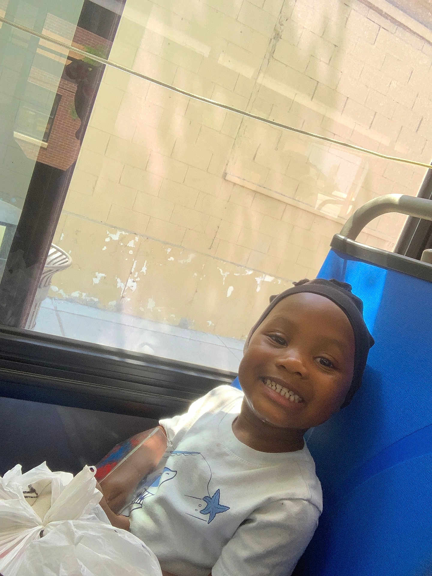 Aijalon is registered to the contest to win money with this photo: child, smiling, boy, seat, window, reflection, cap, shirt, starfish, indoor, daylight, plastic_bag, happy, portrait, casual, candid, sidewalk, wall, blue_seat, person