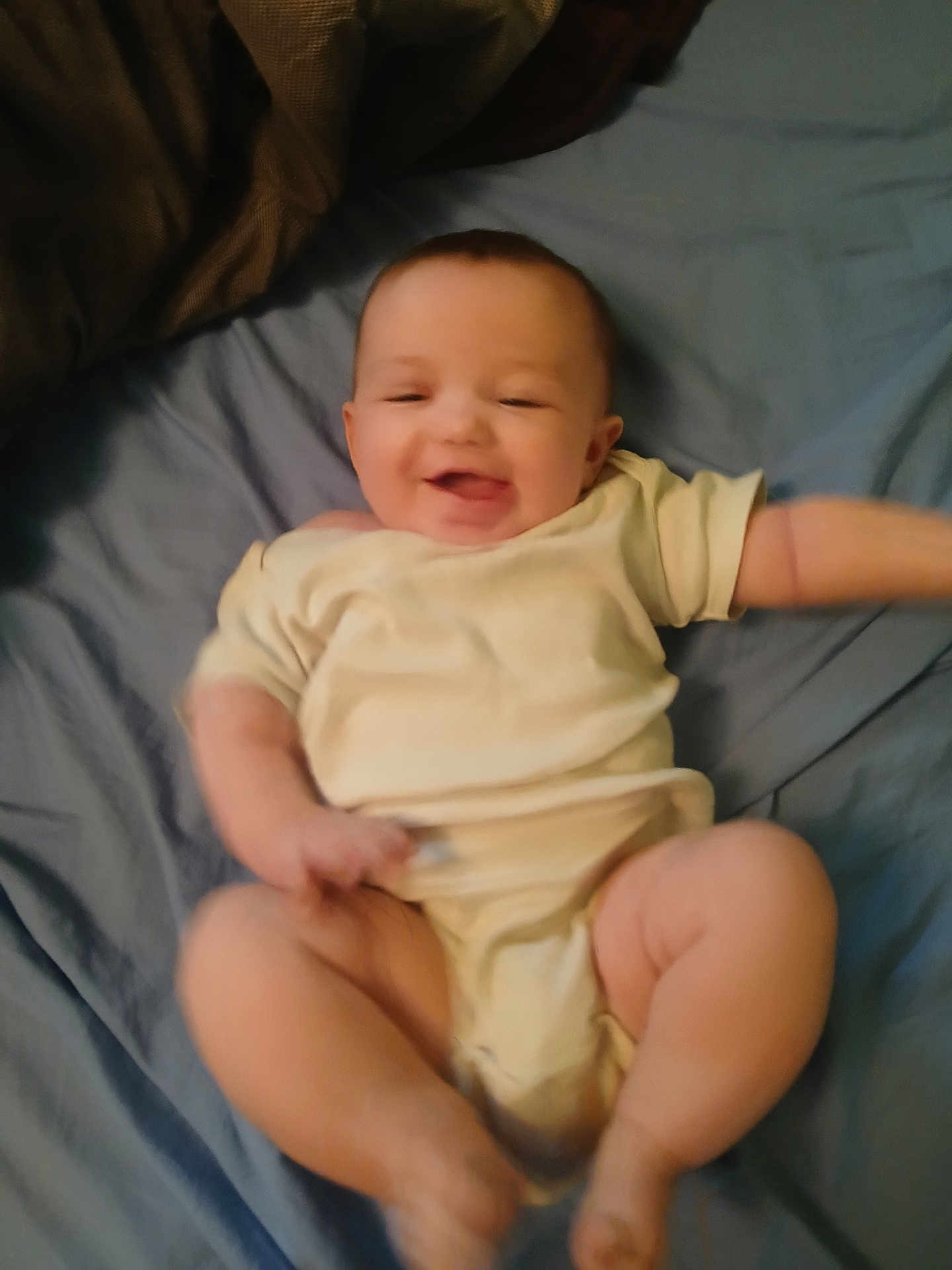 Paige joined the competition — help win amazing prizes! baby, infant, smiling, lying_down, onesie, bed, blue_bedspread, happy, chubby_legs, arms, face, skin, head, cute, child, person, indoor, soft_lighting, motion_blur, playful