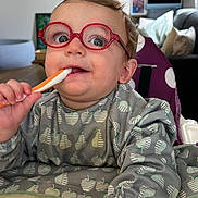 Théo participe au concours pour gagner de l'argent avec cette photo : toddler, child, glasses, spoon, bib, apple_pattern, pear_pattern, high_chair, indoor, living_room, couch, pillow, table, person, face, hand, clothing, cute, baby, feeding
