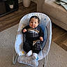 baby, infant, baby_rocker, clothing, white_socks, face, person, carpet, living_room, couch, plant, potted_plant, wood_floor, furniture, indoor, smiling, comfort, cute, home_decor, relaxation