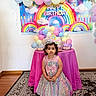 child, birthday, party, balloons, rainbow, tiara, dress, pearls, table, cake, pink, pastel_colors, celebration, floor_rug, indoor, decorations, shoes, curly_hair, young_girl, festive