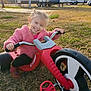child, girl, smiling, tricycle, outdoor, grass, pink_jacket, toy, play, vehicle, happy, daylight, park, footwear, casual_clothing, transportation, nature, recreation, person, leisure