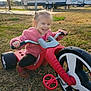 Athena is registered to the contest to win money with this photo: toddler, child, tricycle, pink_jacket, outdoor, grass, smiling, happy, toy, vehicle, play, daylight, parked_trailer, recreational_vehicle, autumn, casual_clothing, fun, young_child, nature, sunlight
