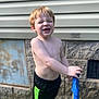 child, boy, shirtless, shorts, outdoor, garden_hose, hose_nozzle, stone_wall, siding, grass, sneakers, sunlight, playful, expression, standing, hand, toy, summer, daytime, casual