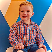 Ryland is registered to the contest to win money with this photo: child, boy, red_hair, plaid_shirt, denim_shorts, smiling, sitting, indoor, geometric_wall, colorful_background, portrait, casual_clothing, happy, cute, young_child, person, fashion, short_hair, bright_colors, playful