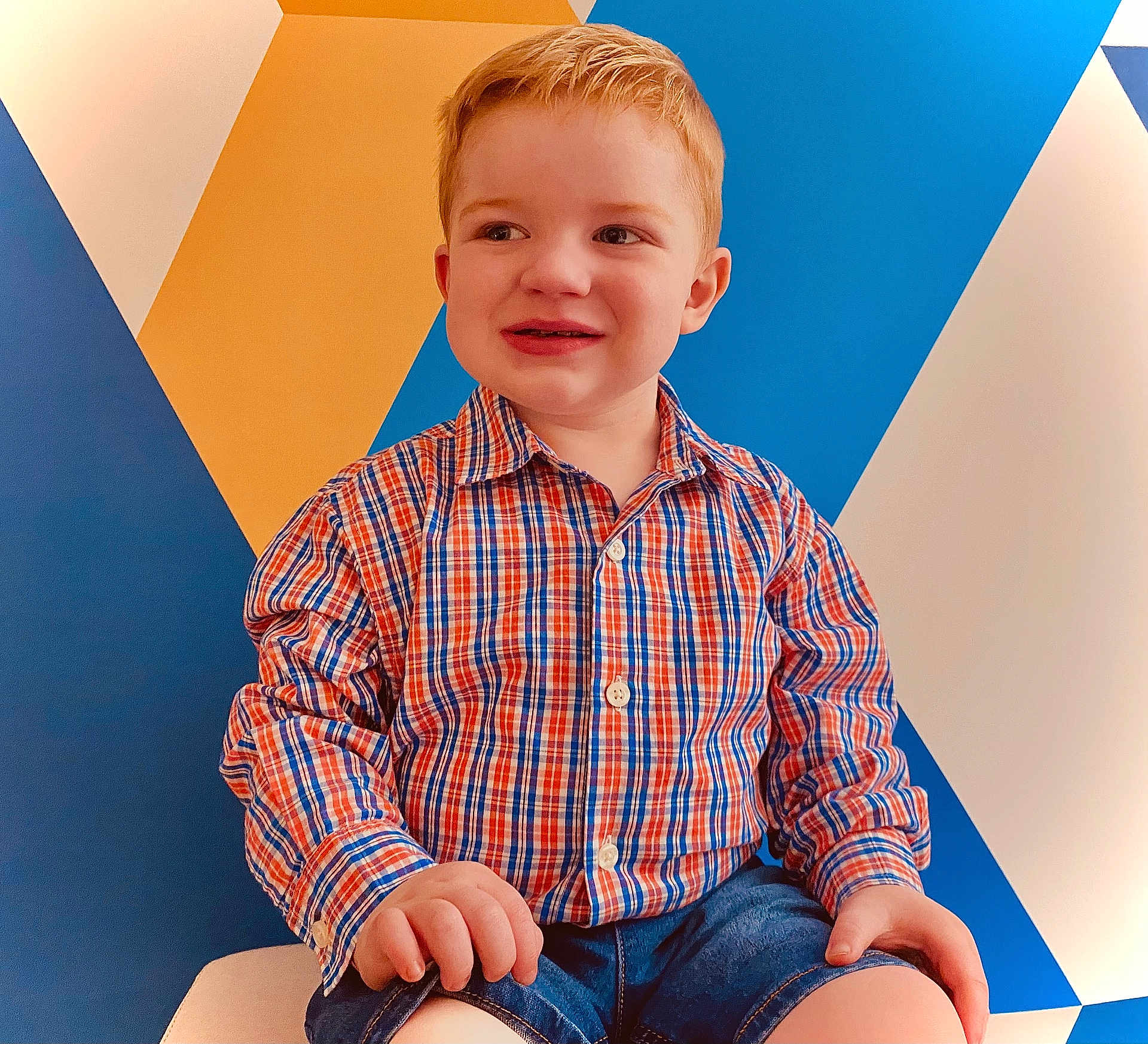 Ryland is registered to the contest to win money with this photo: child, boy, red_hair, plaid_shirt, denim_shorts, smiling, sitting, indoor, geometric_wall, colorful_background, portrait, casual_clothing, happy, cute, young_child, person, fashion, short_hair, bright_colors, playful