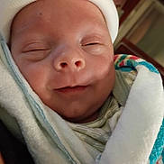 Ashland is registered to the contest to win money with this photo: newborn, baby, infant, swaddled, sleeping, smiling, face, portrait, close_up, white_hat, knit_cap, blanket, colorful_blanket, wrapped, cozy, indoors, lying_down, peaceful, family_photo, baby_face