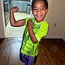 child, boy, smiling, flexing, muscle, green_shirt, purple_shorts, white_shoes, indoor, wooden_floor, kitchen, background_blur, happy, posing, short_hair, casual_clothing, person, playful, confident, standing