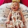 Presley is registered to the contest to win money with this photo: baby, smiling, laughing, headband, dress, cushion, patterned_clothing, joyful, happy, child, infant, sitting, indoor, red_background, white_cushion, text_print, cute, playful, portrait, closeup