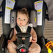 Presley is registered to the contest to win money with this photo: baby, car_seat, child, infant, straps, warning_labels, toy, pink_clothing, blue_eyes, hair, seatbelt, safety, indoors, car_seat_cushion, red_button, curious_expression, skin, foot, hand, closeup
