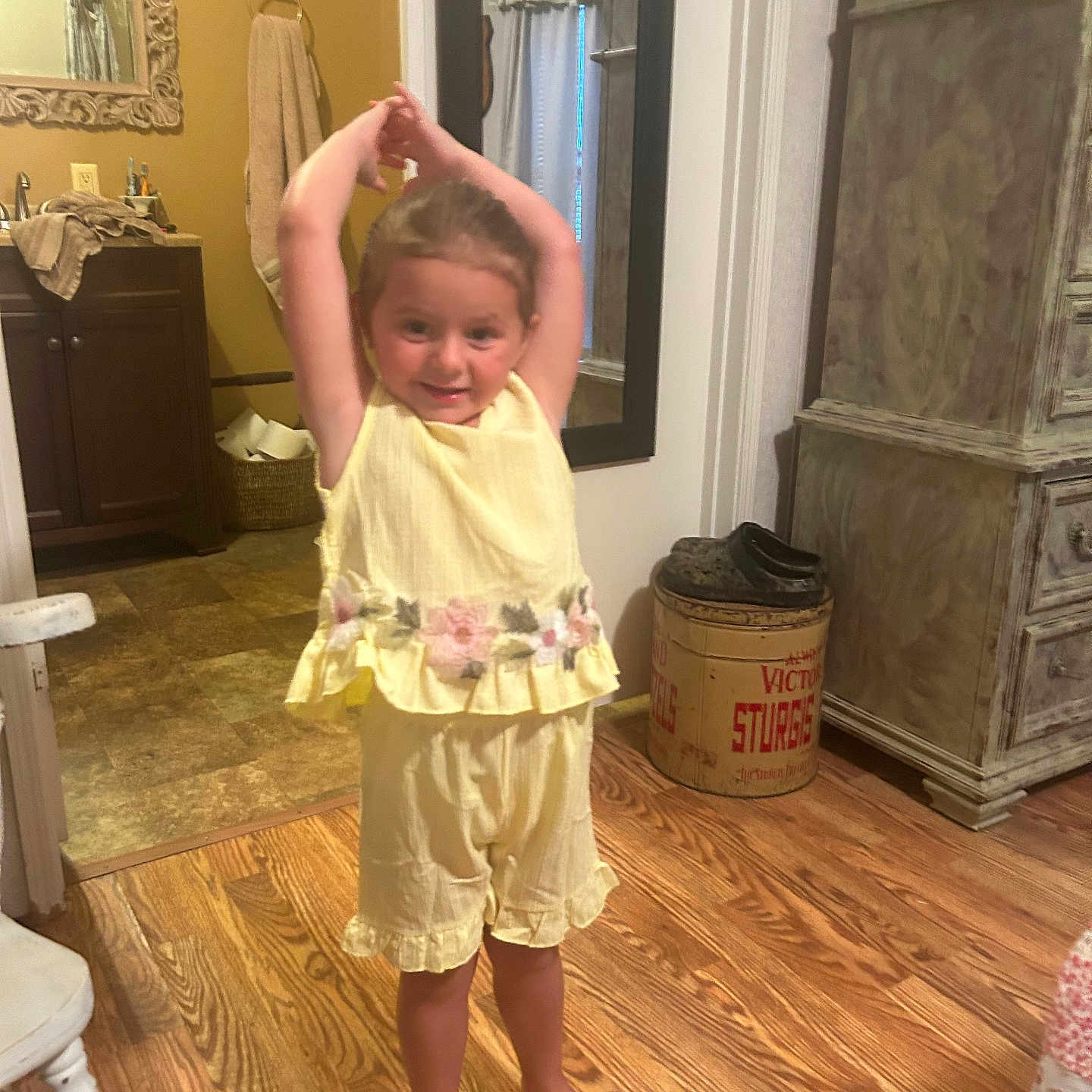 Ella joined the competition — help win amazing prizes! child, girl, toddler, smiling, standing, stretching, arms_up, yellow_outfit, dress, floral_pattern, barefoot, indoors, bathroom, mirror, sink, towel, wooden_floor, furniture, storage_barrel, portrait