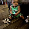 black_and_white_shoes, blurred_background, casual, child, couch, curious_expression, green_clothing, home_interior, indoor, person, relaxed, remote_control, short_hair, sitting, sneakers, sole_of_shoes, sportswear, toddler, wooden_floor, young_child