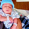 adult_hand, baby, bed, blanket, blue_hat, child, cradling, cute, dinosaur_print, hand, hospital, hospital_wristband, indoor, infant, newborn, onesie, person, portrait, sleepy, wooden_floor