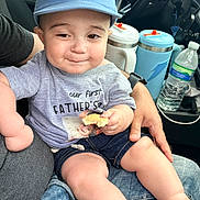 Gabriel is registered to the contest to win money with this photo: baby, child, cap, cookie, denim_shorts, person, car_interior, water_bottle, seat, hand, smile, toddler, clothing, sitting, lap, cute, face, casual, food, parent