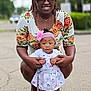 woman, baby, child, outdoor, street, sunglasses, headband, floral_dress, pink, smile, holding_hands, footwear, traffic_lights, pavement, trees, necklace, person, summer, cute, happy