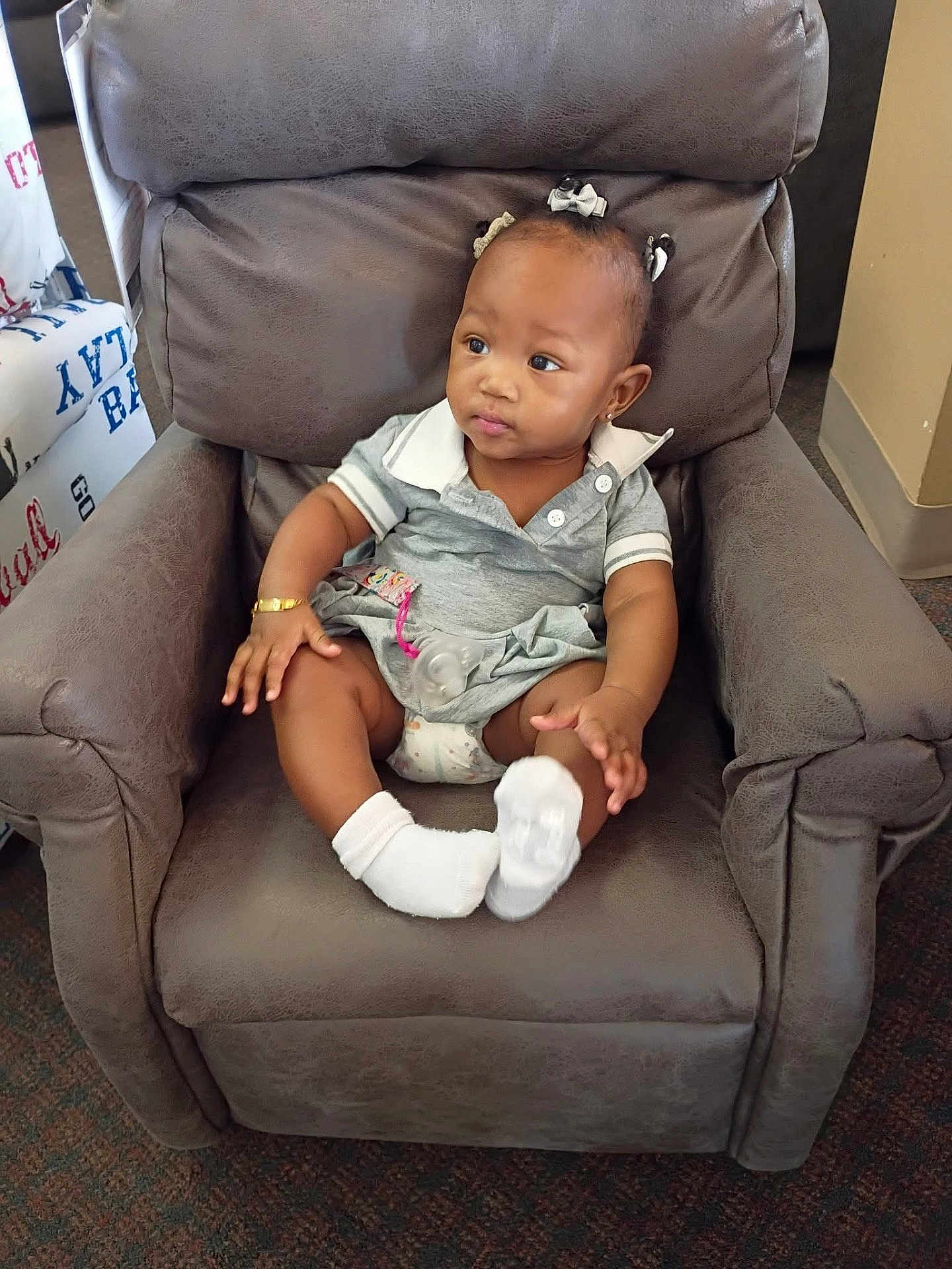 Lyndon is registered to the contest to win money with this photo: toddler, child, baby, armchair, grey_clothing, white_socks, hair_bows, indoor, carpet, bracelet, pacifier, curious_expression, seated, furniture, living_room, small_hands, skin, face, cute, portrait
