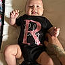 Riley joined the competition — help win amazing prizes! baby, infant, onesie, black_clothing, pink_text, tattoo, hand, cushion, lying_down, skin, chubby_cheeks, happy, indoor, person, child, cute, smiling, arm, leg, portrait