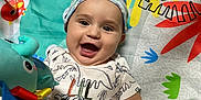 Orion is registered to the contest to win money with this photo: baby, smiling, dinosaur_onesie, hat, play_mat, toy, plush_toy, colorful, infant, happy, child, lying_down, cute, patterned_clothing, cheerful, indoors, playtime, bright_colors, closeup, hand