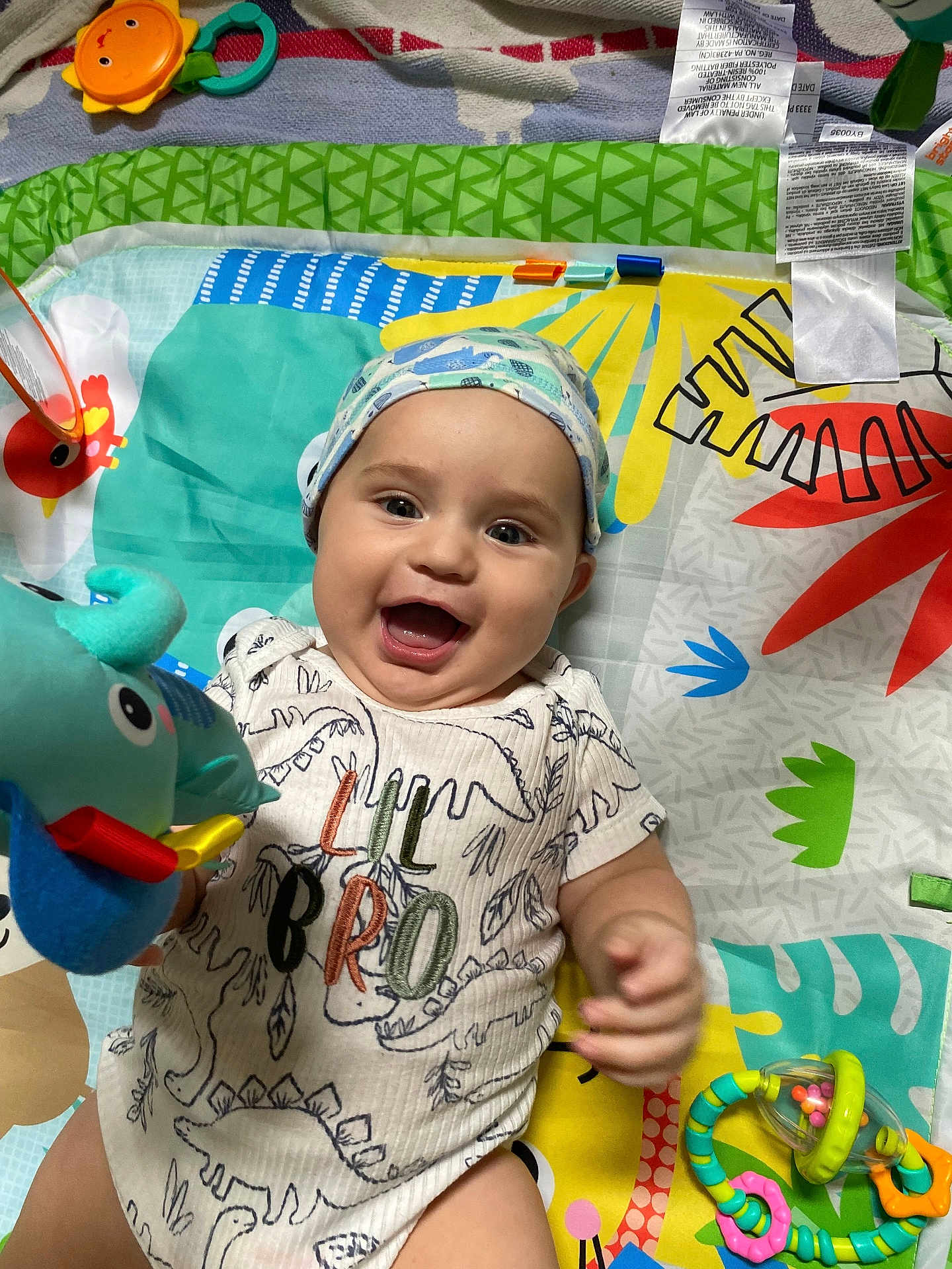 Orion is registered to the contest to win money with this photo: baby, smiling, dinosaur_onesie, hat, play_mat, toy, plush_toy, colorful, infant, happy, child, lying_down, cute, patterned_clothing, cheerful, indoors, playtime, bright_colors, closeup, hand