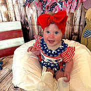Blakely is registered to the contest to win money with this photo: baby, child, smiling, red_bow, patriotic, american_flag, garland, wooden_background, cushion, white_socks, blue_eyes, cute, infant, sitting, clothing, decor, happy, portrait, indoor, celebration
