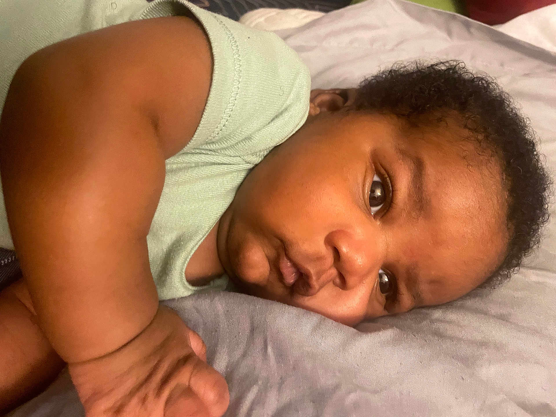 Adonis is registered to the contest to win money with this photo: baby, infant, face, arm, hand, pillow, bedding, onesie, closeup, curly_hair, eye, cheek, nose, laying_down, skin, expression, portrait, sleepy, cute, indoors