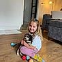 Reggie joined the competition — help win amazing prizes! child, dog, puppy, indoor, floor, toy, tennis_ball, socks, smiling, hair, couch, lamp, door, wood_floor, white_shirt, pink_pants, yellow_socks, happy, hug, living_room
