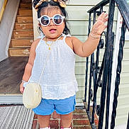 Sahirah is registered to the contest to win money with this photo: toddler, child, sunglasses, hair_buns, white_top, blue_shorts, white_sneakers, bracelets, necklace, purse, porch, brick_floor, doorway, railing, hand_raised, outdoor, fashion, cute, standing, smiling