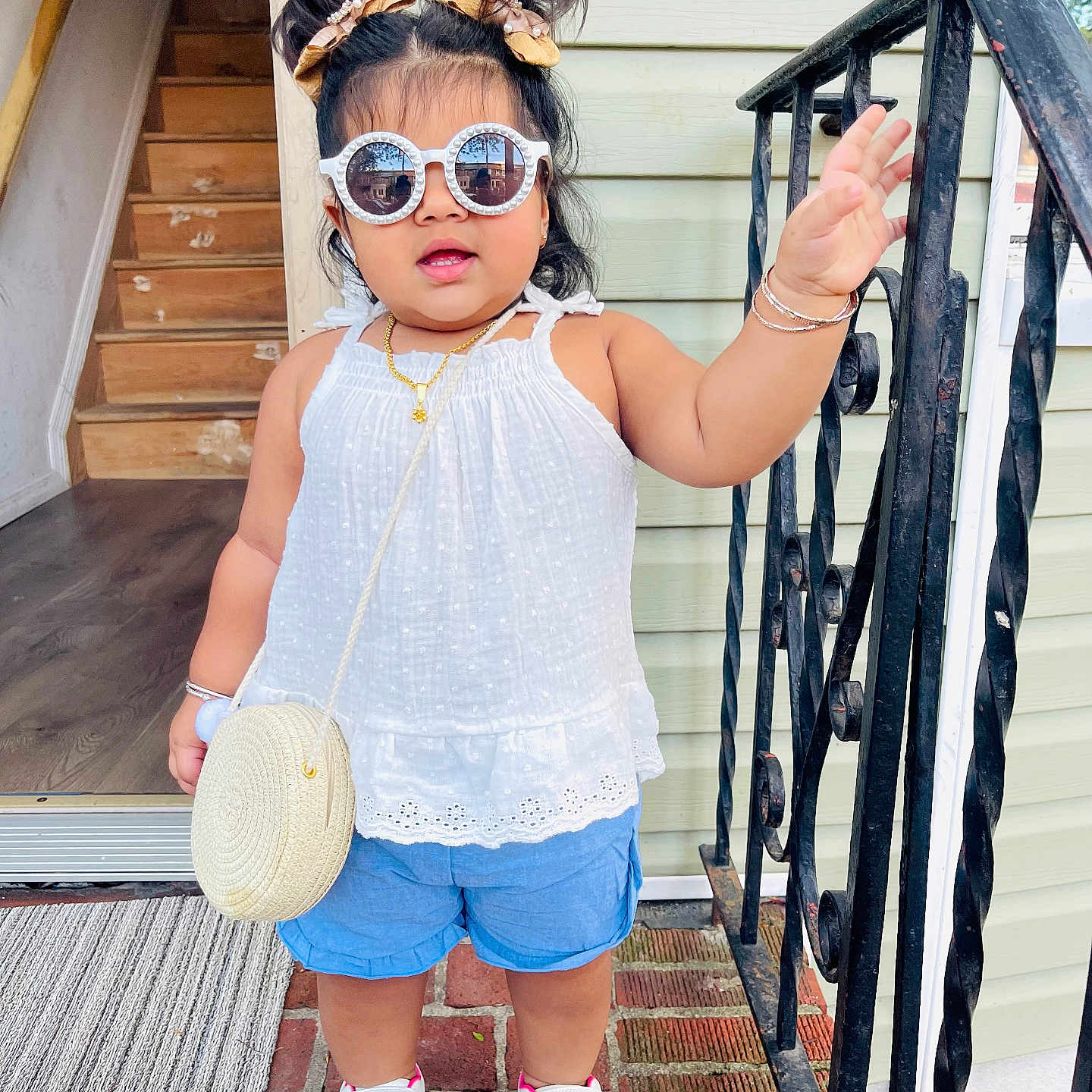 Sahirah is registered to the contest to win money with this photo: blue_shorts, bracelets, brick_floor, child, cute, doorway, fashion, hair_buns, hand_raised, necklace, outdoor, porch, purse, railing, smiling, standing, sunglasses, toddler, white_sneakers, white_top