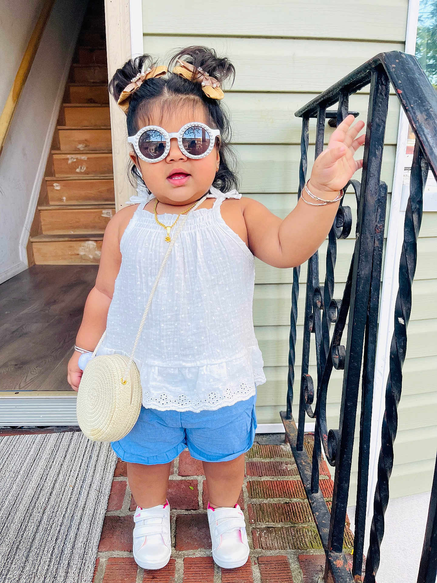 Sahirah is registered to the contest to win money with this photo: toddler, child, sunglasses, hair_buns, white_top, blue_shorts, white_sneakers, bracelets, necklace, purse, porch, brick_floor, doorway, railing, hand_raised, outdoor, fashion, cute, standing, smiling