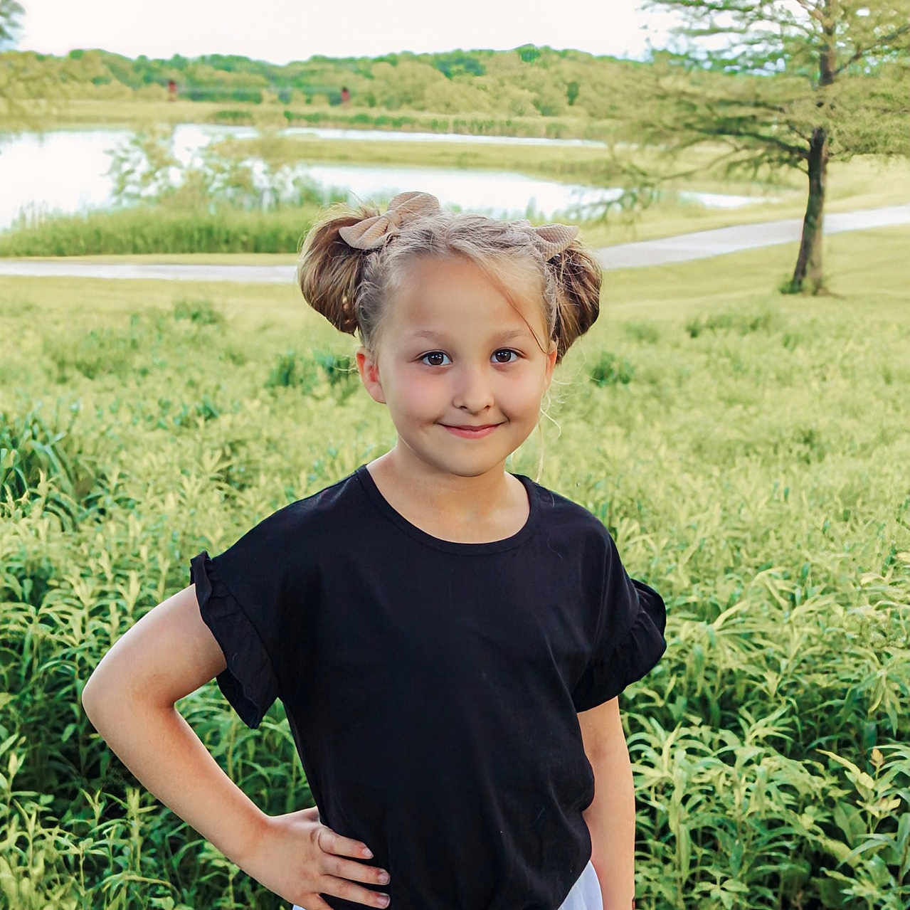 Callie is registered to the contest to win money with this photo: black_shirt, bows, child, field, girl, grass, greenery, hair_buns, happy, lake, nature, outdoor, portrait, posing, smile, summer, sunlight, trees, white_skirt, young