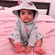 Ajooni is registered to the contest to win money with this photo: baby, child, hoodie, grey_clothing, pink_bedspread, bed, indoor, cute, smiling, sitting, infant, person, face, hands, feet, headwear, cozy, young_child, expression, room