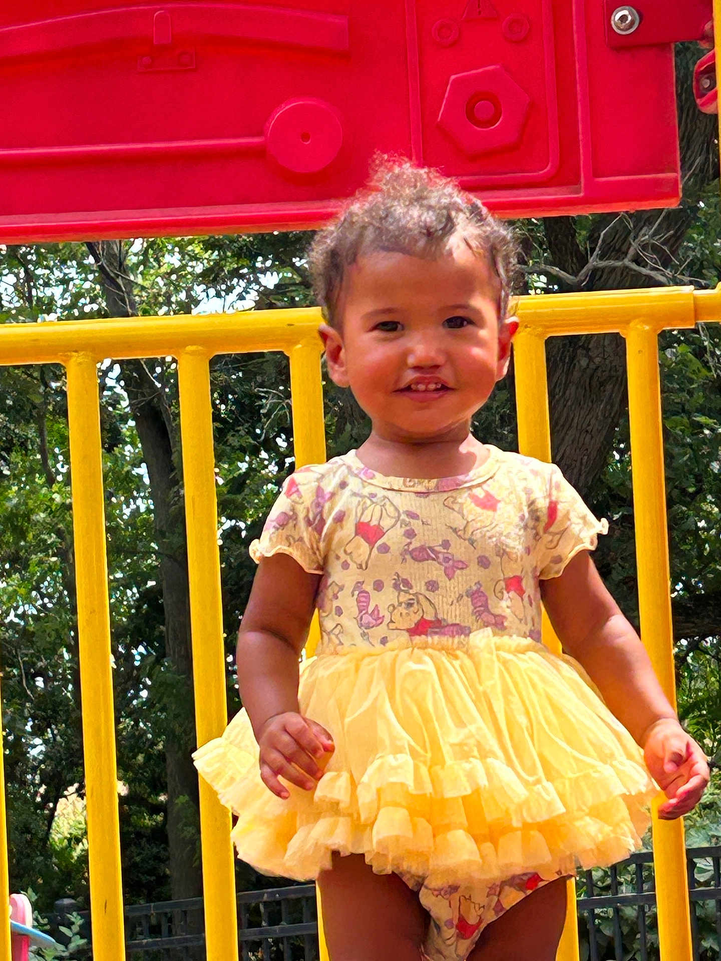 Davina is registered to the contest to win money with this photo: toddler, child, yellow_dress, playground, outdoor, smiling, trees, red_panel, yellow_bars, sunlight, cute, happy, person, summer, nature, park, curly_hair, standing, daytime, playtime
