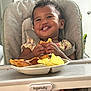 Davina is registered to the contest to win money with this photo: toddler, child, high_chair, food, snacks, smile, happy, meal, indoor, feeding, plate, hands, curly_hair, baby_clothes, table, feet, seatbelt, person, face, cute