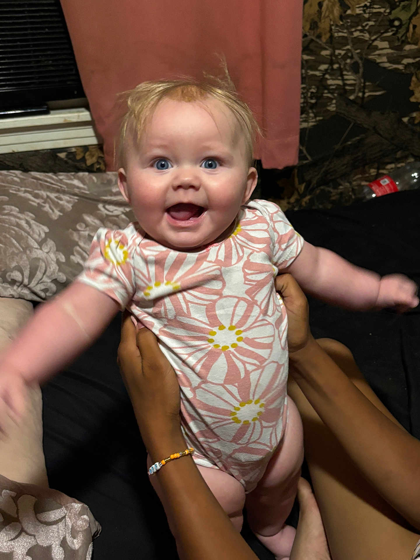 Indie is registered to the contest to win money with this photo: baby, infant, child, smiling, happy, blue_eyes, floral_onesie, arms_outstretched, hands_holding, indoor, bed, curtain, blanket, person, skin, joy, cute, young_child, human, portrait