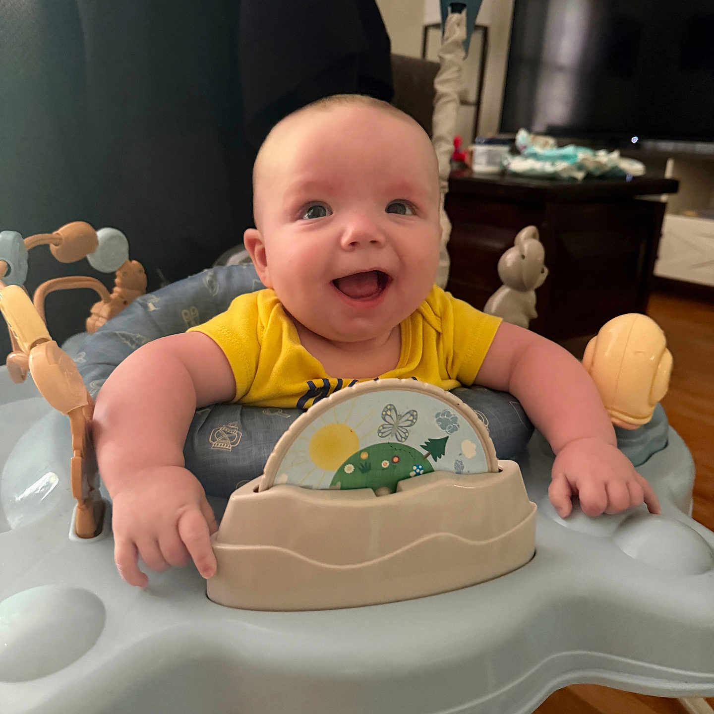 Oliver joined the competition — help win amazing prizes! baby, smiling, indoor, yellow_shirt, activity_center, toys, happy, infant, furniture, television, wooden_floor, face, person, child, home, cute, plush_toy, seat, expression, bright