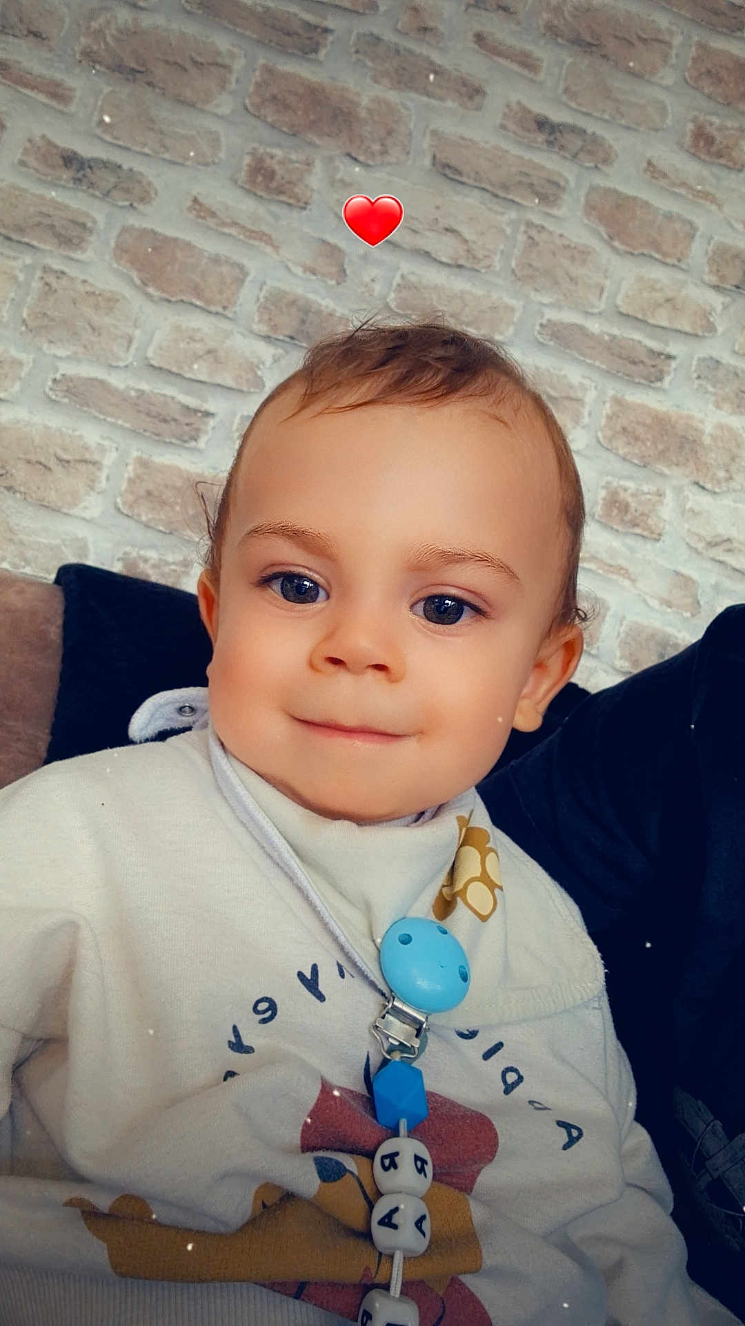 Raphaël a rejoint le concours — aidez-le/la à gagner de superbes lots ! toddler, child, face, smile, pacifier, clothing, indoor, brick_wall, cute, blue_eyes, young_child, portrait, sitting, baby, person, happy, cozy, casual, accessory, heart_emoji