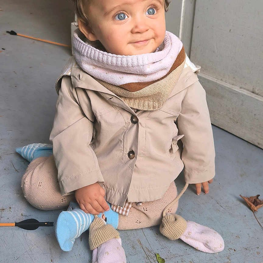 Alix a rejoint le concours — aidez-le/la à gagner de superbes lots ! baby, beige_jacket, bicycle, blue_eyes, child, clothing, concrete, curious, curly_hair, cute, floor, indoor, person, portrait, scarf, sitting, smile, socks, toddler, wall
