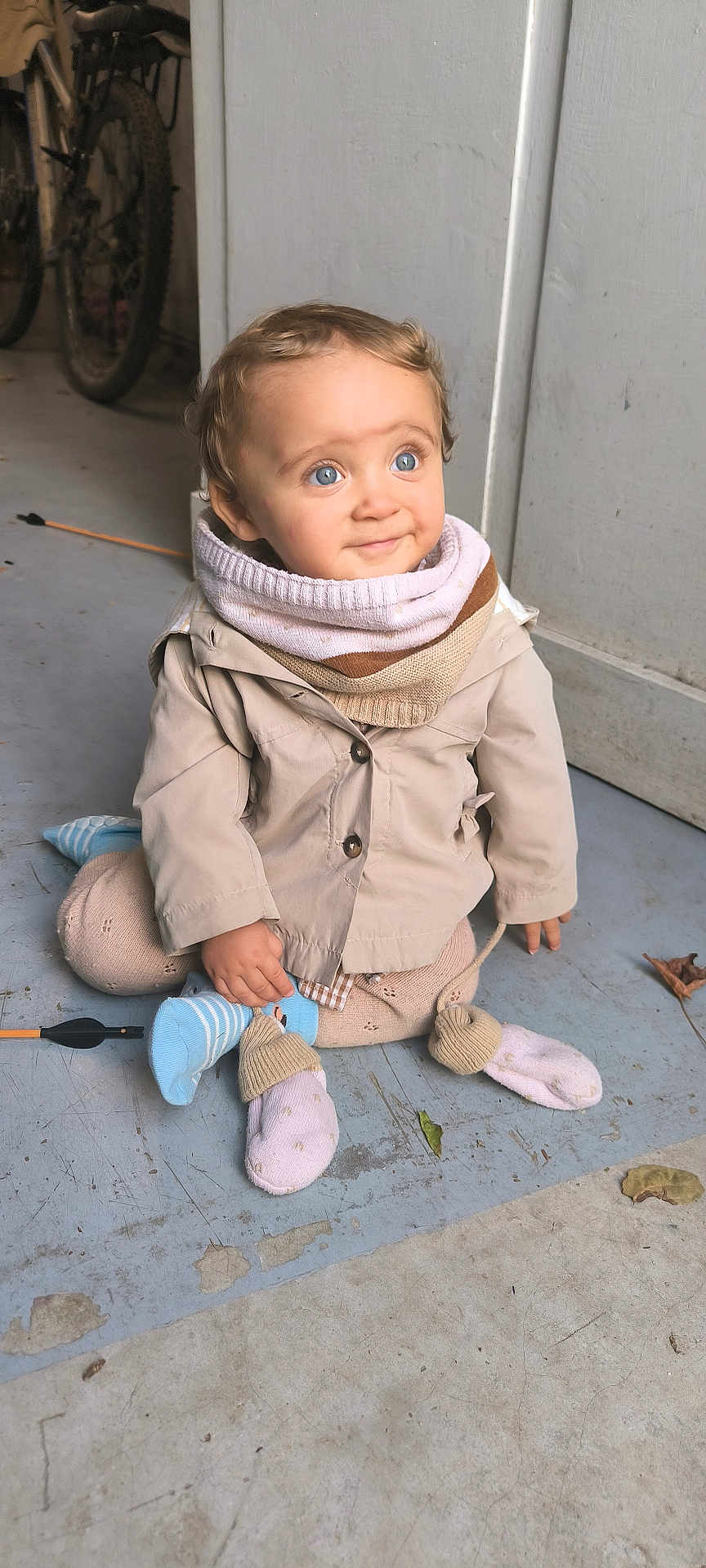 Alix a rejoint le concours — aidez-le/la à gagner de superbes lots ! toddler, child, blue_eyes, curly_hair, scarf, beige_jacket, socks, floor, bicycle, indoor, concrete, wall, sitting, smile, cute, curious, clothing, person, portrait, baby
