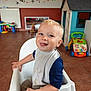baby, bib, child, clothing, colorful, floor, furniture, happy, high_chair, indoor, person, playful, playroom, room, seat, smiling, toddler, tongue_out, toys, wall_decorations