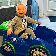 Elyo participe au concours pour gagner de l'argent avec cette photo : blue, child, couch, door, floor, green, grey_outfit, happy, indoor, living_room, mickey_mouse, person, play, seatbelt, smiling, text_on_clothing, toddler, toy_car, wheels, window
