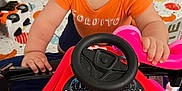 Nathan is registered to the contest to win money with this photo: toddler, child, orange_shirt, toy_car, steering_wheel, pink_toy, play_area, indoor, colorful, plush_toy, floor_mat, black_leather_sofa, baby, cute, curious, playing, hands, face, expression, home
