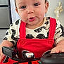 Nathan is registered to the contest to win money with this photo: baby, child, toy_car, red_clothing, steering_wheel, indoor, playful, cute, face, expression, hair, black_and_white_shirt, buttons, hand, furniture, floor, home, person, portrait, infant