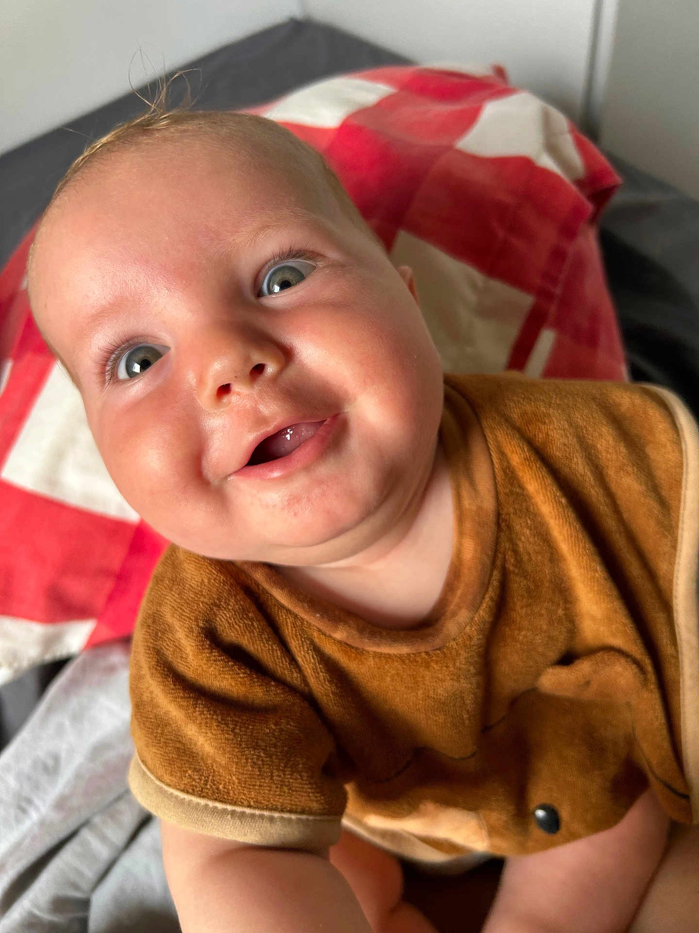 Jules participe au concours pour gagner de l'argent avec cette photo : baby, child, smiling, blue_eyes, brown_clothing, close_up, face, infant, joyful, bed, blanket, red_and_white, cute, skin, happy, portrait, indoor, person, young_child, expression